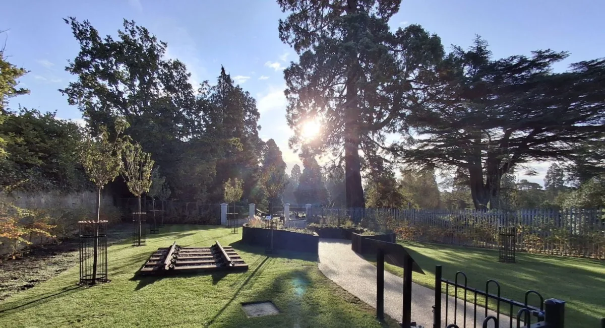 The Brookwood Reflective Garden in autumn sun, with a short section of railway track on the lawn, young trees and a path leading to the cemetery gates