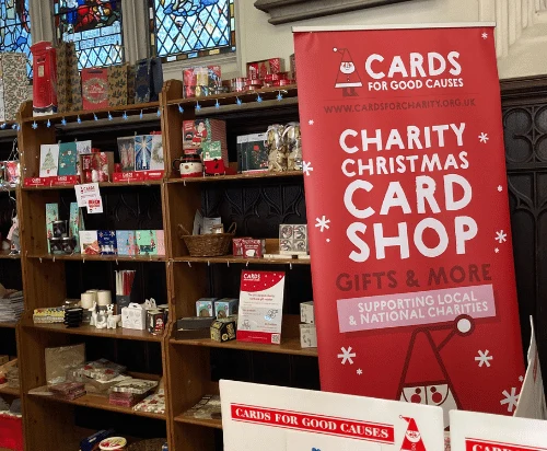 Wooden shelves of Christmas cards and gifts beneath stained glass windows, beside a red Cards for Good Causes Charity Christmas Card Shop banner