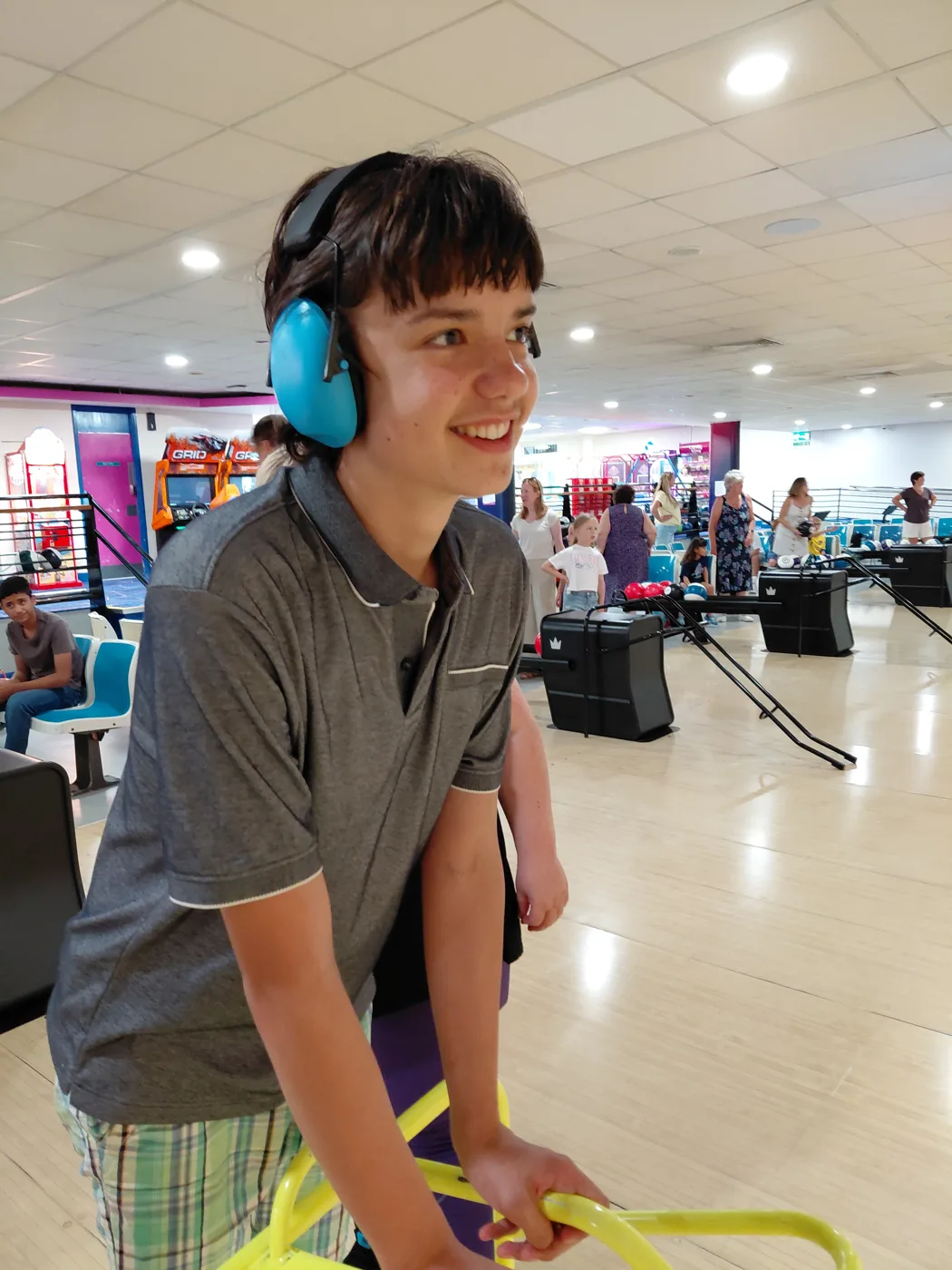 Leo, a teenage boy wearing blue ear defenders, smiling at a bowling alley