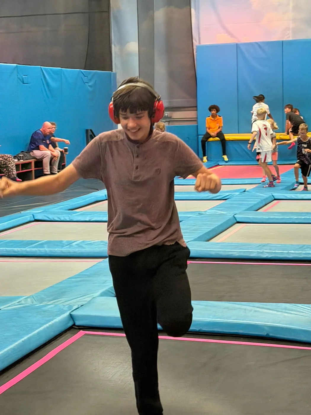Leo, wearing red ear defenders, grinning as he jumps at a trampoline park