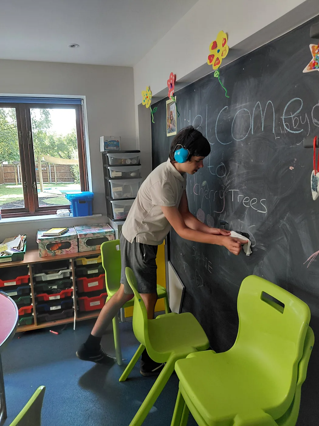 Leo, wearing blue ear defenders, wiping a chalkboard with 'Welcome to Cherry Trees' written on it