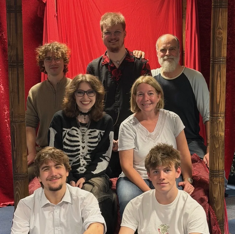 Seven members of the cast of The Lion in Winter posing together in front of a red curtain between two carved wooden bedposts